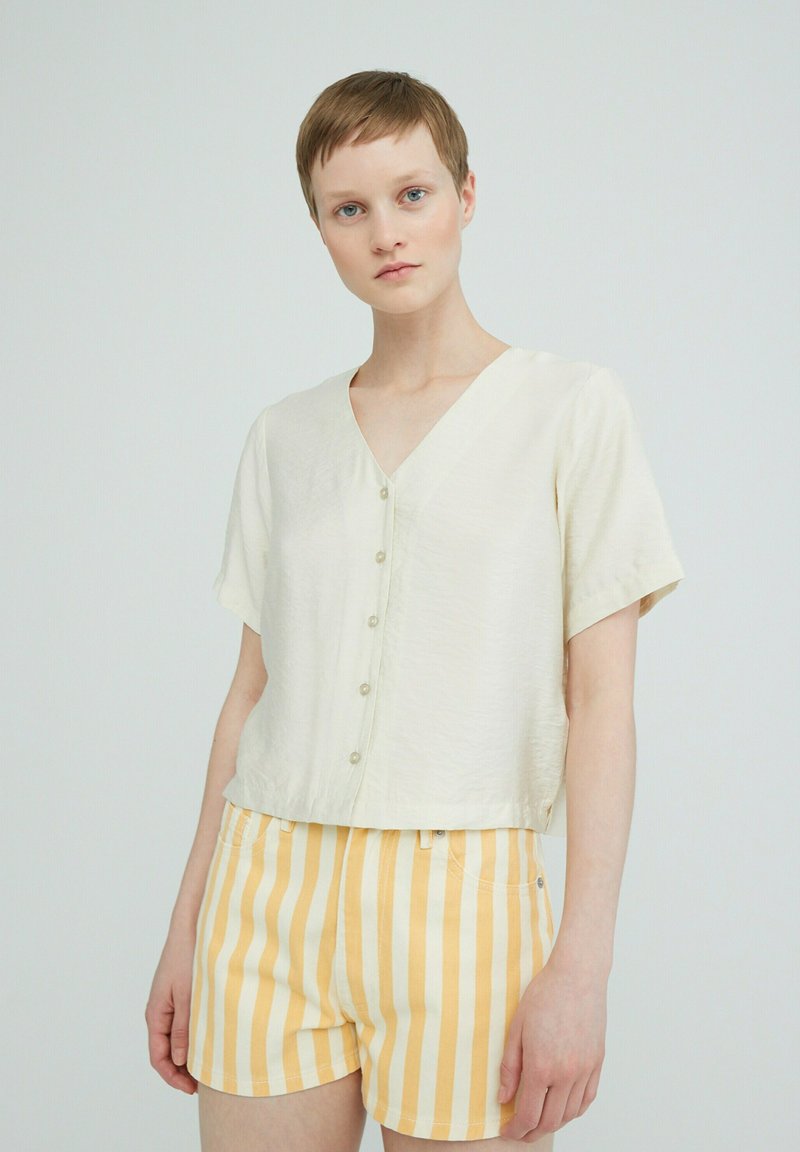 Beige blouse met korte mouwen en v-hals, voorzien van knopen, gecombineerd met gele en witte gestreepte shorts. Zachte stof en ontspannen pasvorm.