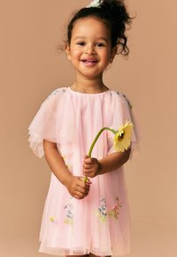 Rosa Tüllkleid mit kurzen, geschichteten Ärmeln, ausgestattet mit buntem Blumenstick. Das Kind hält eine gelbe Blume. Einfacher runder Ausschnitt.