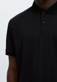Persona che indossa una polo nera semplice a maniche corte con collo e due bottoni visibili sulla patta.