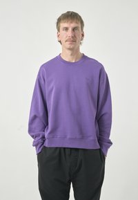 Purple sweatshirt with a round neck and ribbed hem, featuring long sleeves and a small logo on the left chest. Paired with black pants.