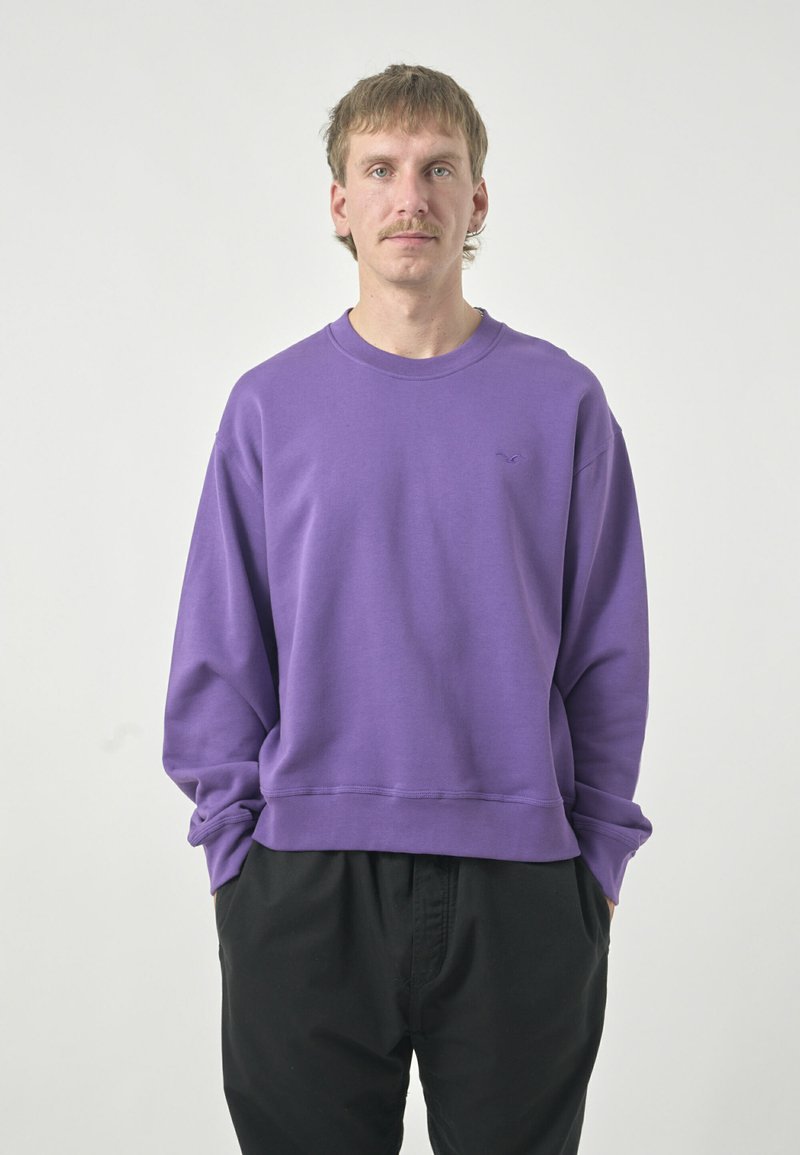 Purple sweatshirt with a round neck and ribbed hem, featuring long sleeves and a small logo on the left chest. Paired with black pants.