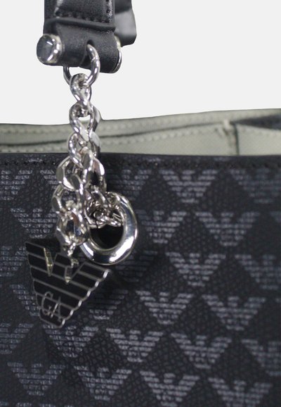 Schwarze Handtasche mit einem strukturierten Muster, silbernem Kettendetail und einem Logo-Anhänger. Der glatte Ledertragegurt verbindet sich mit dem oberen Teil der Tasche.