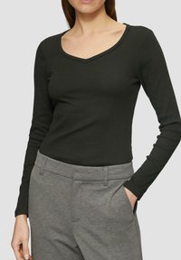 Schwarzes langärmliges Oberteil mit V-Ausschnitt, aus geripptem Material. Kombiniert mit grauen Hosen mit flachem Bund und Seitentaschen.