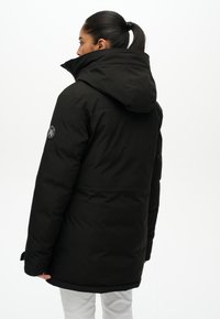 Czarna ocieplana kurtka z dużym kapturem, pagonami na ramionach i okrągłą naszywką z logo po lewej stronie. Materiał wydaje się gładki i matowy.