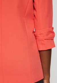 Blusa coral con un corte entallado, que presenta un cuello con pico y mangas enrolladas. Textura suave con detalles de costura sutiles a lo largo de las costuras.