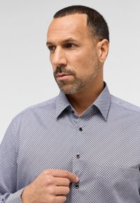 Overhemd met knopen, versierd met een geometrisch patroon in blauw en wit, met donkere knopen. De kraag is gestuctureerd; de stof lijkt glad en textuur te hebben.