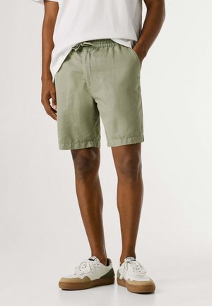 Hombre con pantalones cortos verde claro con cordón, camiseta blanca y zapatillas blancas con suelas marrones, de pie con una mano en el bolsillo.