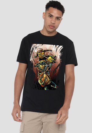 Young man wearing a black t-shirt with colorful Teenage Mutant Ninja Turtles in action poses on the front, beige cargo shorts, neutral background.