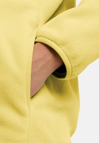 Maglia in felpa gialla con polsino a coste e tasca laterale. Il tessuto è morbido e testurizzato, con una finitura liscia sulla superficie esterna.