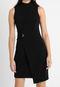 Černé šaty bez rukávů s falešným rolákem, asymetrickým zavinovacím designem, boční sponou a hladkou elastickou látkou.
