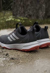 Paire de chaussures de trail noires et grises avec semelles intermédiaires blanches et semelles extérieures rouges sur un sentier de terre près de la verdure.