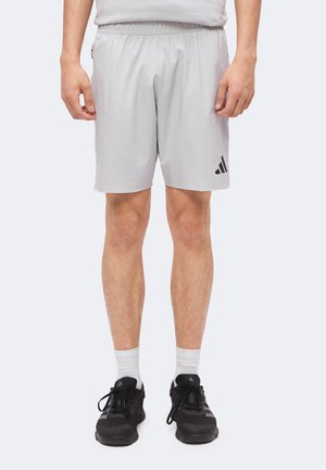Uomo che indossa pantaloncini Adidas grigio chiaro, calzini bianchi e scarpe da corsa Adidas nere, in piedi davanti a uno sfondo semplice.