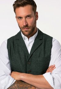 Muž s plnovousem a vyhrnutými rukávy bílé košile, oblečený v tmavě zelené vlněné vestě, se založenýma rukama, na předloktí má tetování.