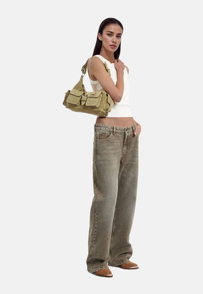 Borsa beige con multiple tasche, accessoriata con una clip d'argento. Il modello indossa una maglia bianca aderente e jeans in denim chiaro e larghi.