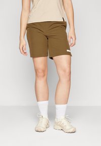 Olivenfargede treningsshorts med en glatt tekstur, med en sidelomme og et lite logopatch. Kombinert med beige joggesko og hvite sokker.
