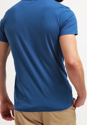 Homme portant un polo bleu uni à manches courtes et un short beige, vu de dos sur un fond uni clair.