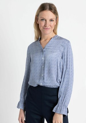 Chemisier bleu léger avec un motif géométrique, devant à boutons et poignets froncés, associé à un pantalon foncé ajusté.