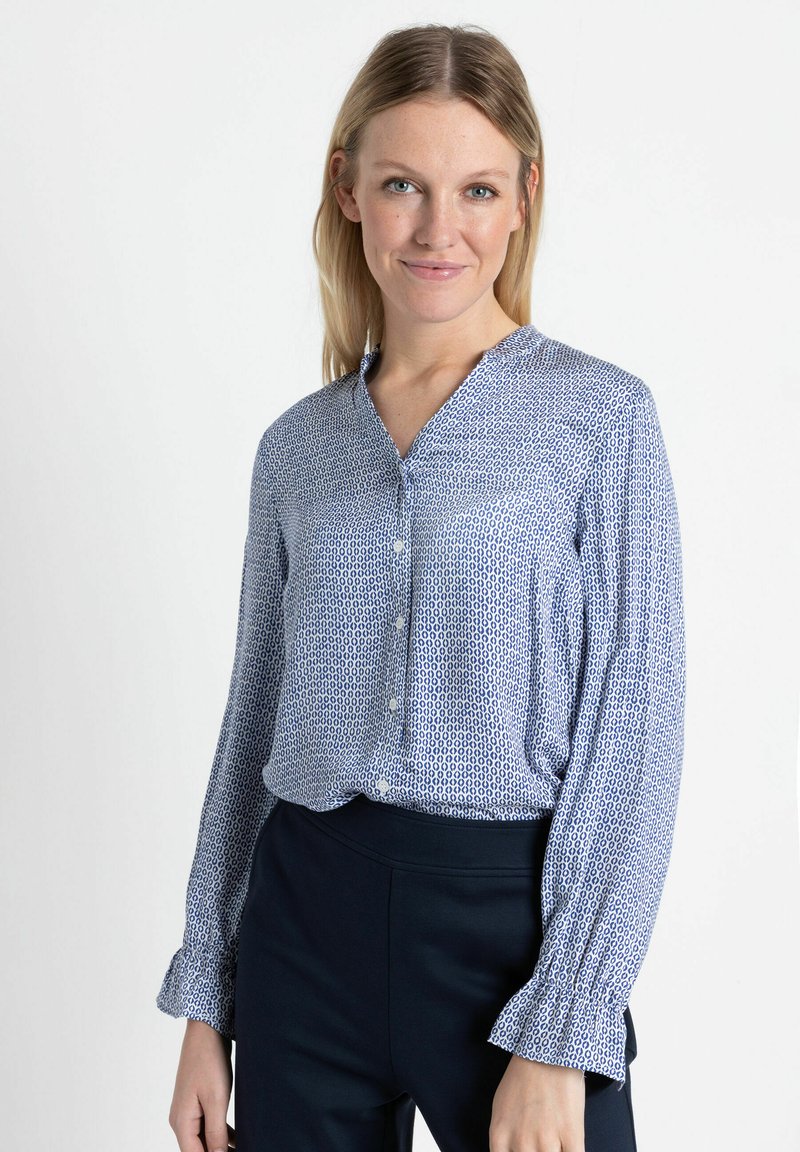 Lightweight blue blouse with a geometric pattern, button-down front, and ruffled cuffs, paired with dark fitted pants.
