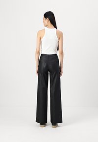 Un débardeur blanc sans manches associé à un pantalon noir à taille haute et jambes larges, mettant en avant une texture lisse et deux poches à l'arrière. Le modèle a les cheveux longs.