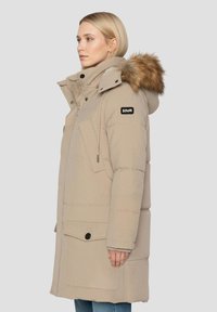 Beżowa pikowana parka z futrzanym kapturem, zamkiem błyskawicznym z przodu i guzikami oraz dużymi bocznymi kieszeniami. Wytrzymały materiał i pikowana faktura. Widoczne logo marki.