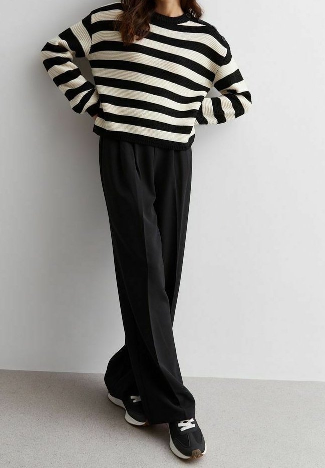 Personne portant un pull à rayures horizontales noires et blanches, un pantalon noir ample et des baskets noires, debout les mains sur les hanches.