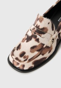 Mocassino con motivo maculato marrone e bianco, con suola nera e punta arrotondata, che mostra la parte superiore e una parte della fodera interna.
