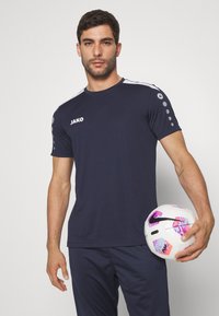 Maglietta sportiva navy a maniche corte, con dettagli bianchi e bottoni grigio scuro sulle spalle. Il modello tiene in mano un pallone da calcio decorato.