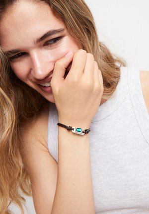 Bracelet en cuir avec un accent rectangulaire en argent et une pierre turquoise. Texture lisse et design ajustable, porté sur une manche gris clair.