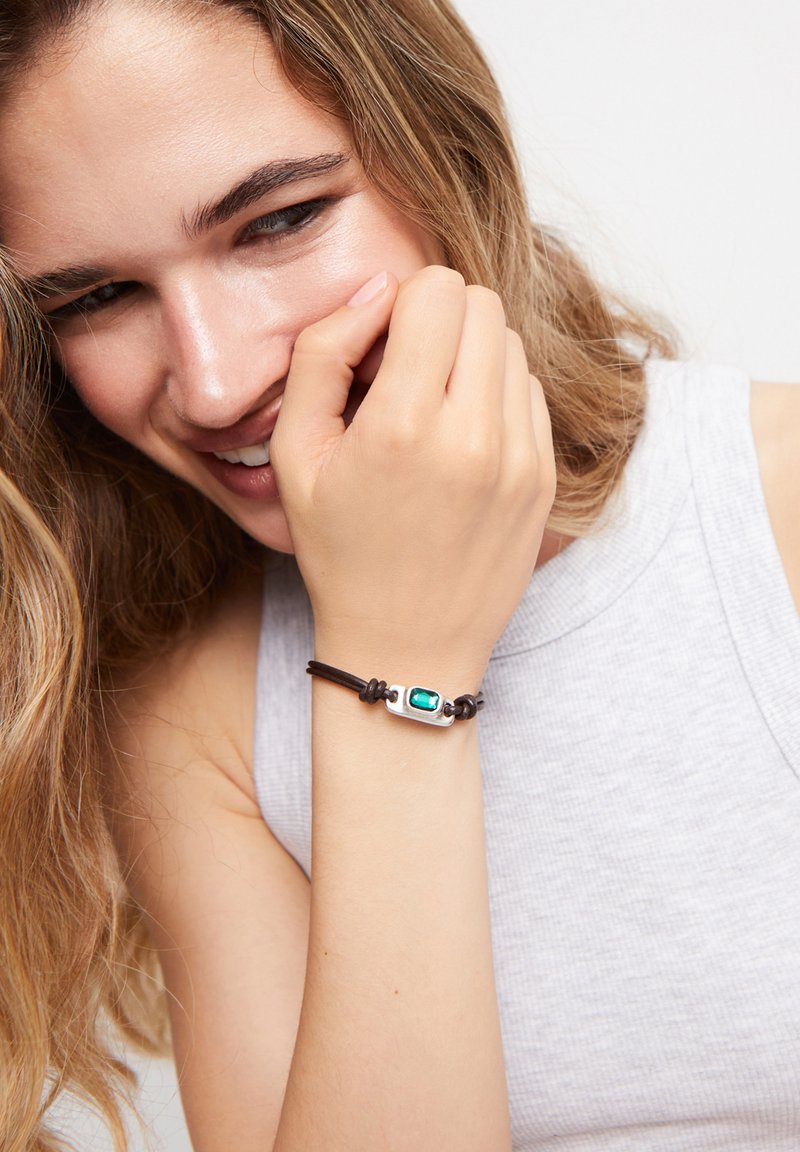 Bracelet en cuir avec un accent rectangulaire en argent et une pierre turquoise. Texture lisse et design ajustable, porté sur une manche gris clair.