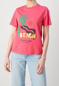 Camiseta de algodón rosa con un diseño gráfico que incluye una palmera, un sol y el texto "BEACH" en amarillo y negro, combinada con vaqueros de color azul claro.