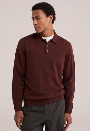Homme portant un polo à manches longues rouge foncé par-dessus un maillot blanc et un pantalon gris foncé, regardant vers sa gauche.