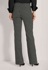 Vrouw in zwart-witte, geometrisch patroon wijde broek en zwarte hakken, gezien vanaf de achterkant tegen een effen achtergrond.