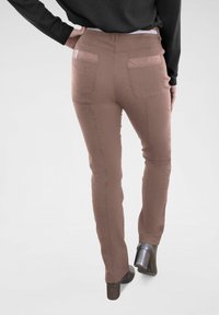 Braune, enganliegende Hose mit glatter Textur, ausgestattet mit Gesäßtaschen und schlankem Design. Getragen mit einem schwarzen Oberteil und Schuhen mit Absatz.