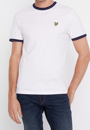 Homme portant un t-shirt blanc à manches courtes avec col et bords de manches bleu marine, petit logo d'oiseau jaune sur la poitrine, associé à un jean bleu foncé.