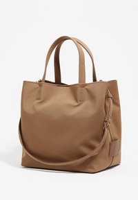 Brązowa torba typu tote z podwójnymi uchwytami, o gładkiej fakturze, zaokrąglonym kształcie i bocznej ozdobnej metce. Minimalistyczny design, zamek błyskawiczny.