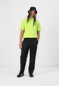 Neon grønn t-skjorte med korte ermer og logo; svarte, løstsittende bukser; svart bøttehatt; svarte joggesko med hvite striper.