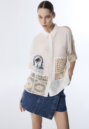 Camisa blanca de botones con paneles de ganchillo, que presenta un diseño bordado de palma. Fabricada con tejido ligero y ajuste relajado. Falda de mezclilla.