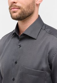 Man met een korte baard, gekleed in een donkergrijze overhemd met knopen, versierd met een klein ruitjespatroon en een borstzak.