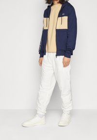 Felpa con zip blu marina con pannelli beige sul petto, tasche a marsupio e pantaloni della tuta bianchi. Sneakers bianche completano l'outfit.