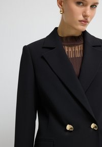 Czarna dwurzędowa marynarka z klapami, złotymi guzikami i gładkim, ustabilizowanym materiałem. Posiada warstwowy brązowy top pod spodem.