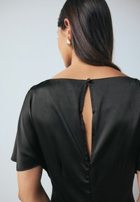 Fekete, rövid ujjú ruha texturált anyagból, kulcslyuk hátkivágással, amelyet egy gomb biztosít; a egyszerűséget és eleganciát hangsúlyozza.