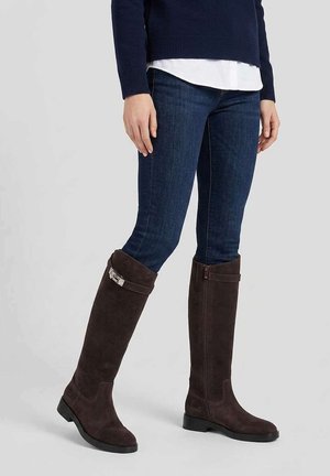 Personne portant un jean skinny bleu foncé et de hautes bottes en daim marron foncé, debout sur un fond clair.