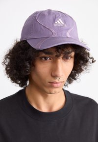 Purple cotton cap with a curved brim, featuring white Adidas logo and stitching accents. Black sweatshirt visible underneath.