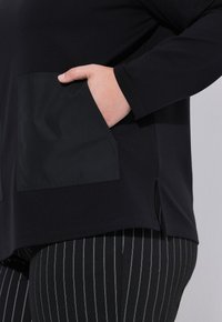 Haut noir à manches longues avec une poche à patch texturée, des fentes sur les côtés et un tissu lisse, associé à un pantalon noir avec des rayures verticales blanches.