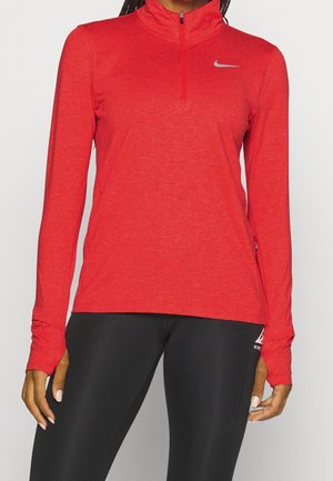 Femme portant un haut sportif rouge à manches longues avec une fermeture éclair au quart et un legging noir, debout devant un fond uni.