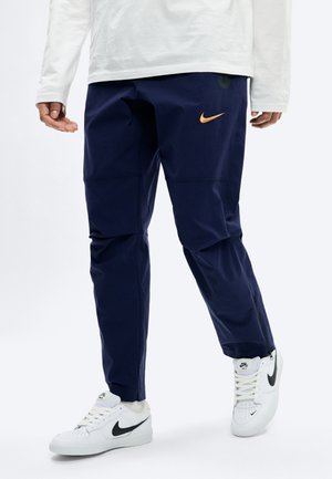 Person iført marineblå Nike-bukser med oransje logo og hvite Nike-sneakers med svart swoosh, stående mot en hvit bakgrunn.