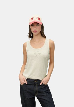 Donna che indossa una canotta bianca, jeans scuri e un cappellino da baseball beige e rosa con testo rosso "1936", mani in tasca, in piedi davanti a uno sfondo uniforme.