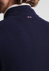 Prenda de punto en azul marino con una textura suave, que presenta un cuello en un suave gris claro y un pequeño detalle bordado en tri-color en la parte posterior.