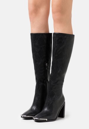 Zwarte kniehoge laarzen met een gestructureerd patroon, spits toelopende neuzen met metalen punten, een zijrits en blokhakken, gedragen op blote benen.