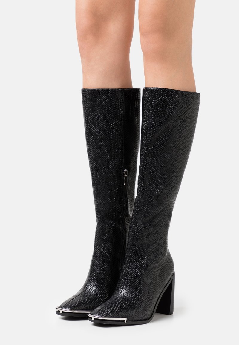 Bottes noires montantes jusqu'aux genoux avec un motif texturé, bouts pointus avec embouts métalliques, fermeture éclair latérale et talons blocs portées sur jambes nues.